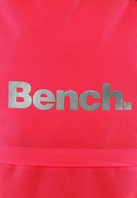Lyst pink stof med en glat tekstur har ordet "Bench." i store, metalliske sølvbogstaver. Enkel, moderne design.