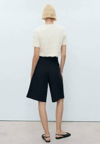 Haut court blanc et duveteux associé à un short bleu marine taille haute. Le mannequin porte des sandales plates noires. Le fond est gris clair.