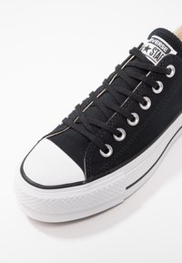Baskets Converse Chuck Taylor noires avec un capuchon en caoutchouc blanc, une semelle intermédiaire blanche et des lacets noirs sur un fond blanc.