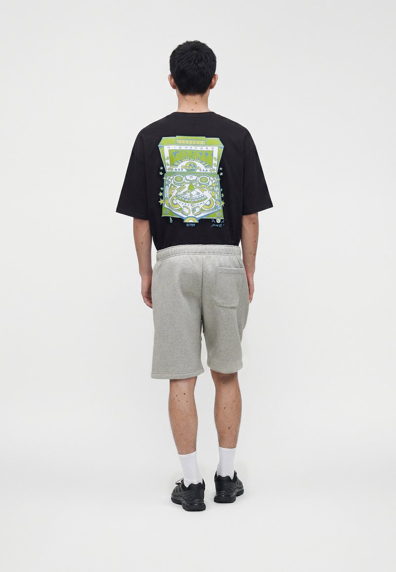 Mann trägt ein schwarzes oversized T-Shirt mit farbenfroher Flipperautomaten-Grafik auf dem Rücken, graue Shorts, weiße Socken und schwarze Sneakers, schaut nach hinten.
