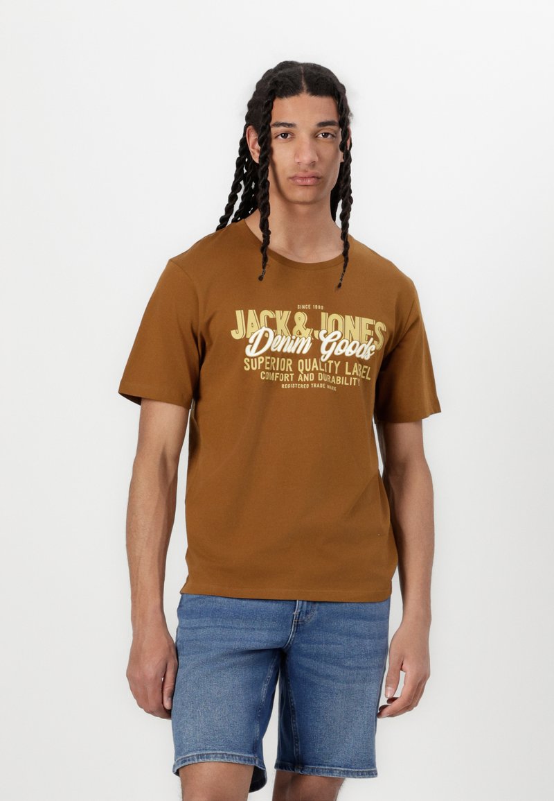 Camiseta de algodón marrón de manga corta, con texto y gráficos en blanco. Combinada con pantalones cortos de mezclilla, el fondo es blanco liso.