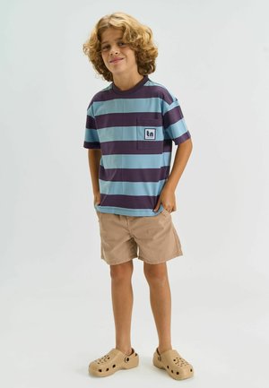 Fiatal, göndör hajú fiú mosolyogva áll, kék és sötét lila csíkos pólót, bézs rövidnadrágot és bézs csúszásmentes cipőt visel.