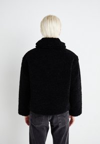 Person with short blond hair facing away, wearing a black textured fleece jacket and dark gray jeans against a plain white background.