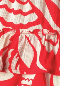 Tissu avec une base blanche texturée et un motif floral abstrait rouge audacieux, comprenant une couture froncée avec un détail de bord volanté.