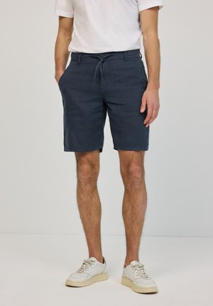 Mann trägt marineblaue Shorts mit Kordelzug, weißes T-Shirt und weiße Sneakers, steht mit den Händen in den Taschen vor einem schlichten Hintergrund.