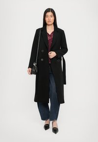 Vrouw met een lange zwarte jas over een bordeaux blouse, blauwe jeans, zwarte puntige schoenen, en een kleine zwarte tas met kettingband.