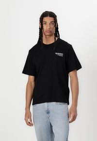 T-shirt in cotone nero con collo rotondo, maniche corte e un piccolo logo bianco sul lato sinistro del petto. Abbinato a jeans denim azzurri.