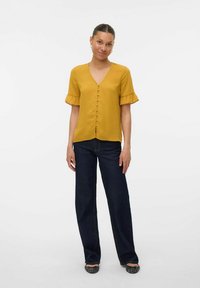 Camicia button-up gialla senape con scollo a V e maniche corte a campana, abbinata a jeans a gamba larga blu scuro e scarpe basse nere.