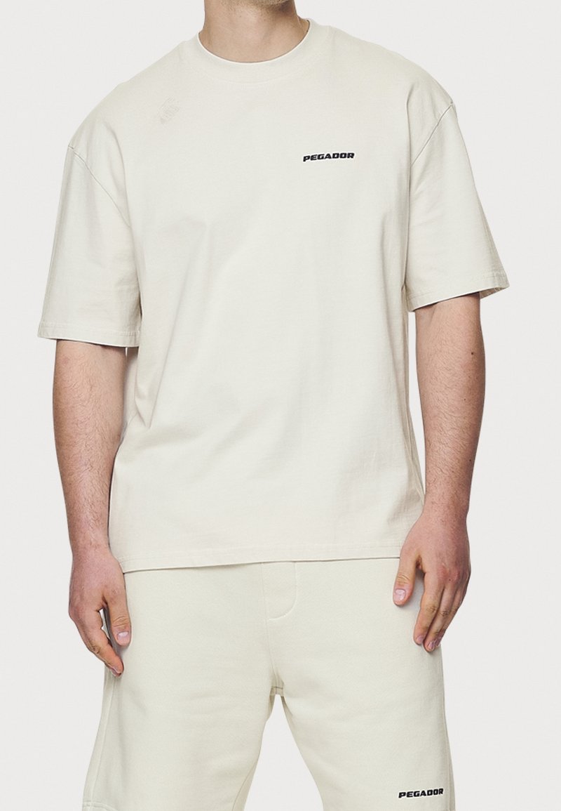 Beige bomullströja med korta ärmar, rund halsringning, med en liten svart "PEGADOR"-logotyp på vänster bröst. Enkel design, avslappnad passform.