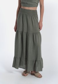 Lunga gonna maxi a strati in lino verde oliva. Caratterizzata da una vita elasticizzata e un orlo arricciato. Abbinata a sandali intrecciati.