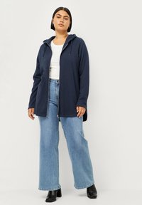 Marineblauwe zip-up hoodie met een trekkoord in de capuchon, gecombineerd met lichtblauwe wijde jeans en zwarte hoge laarzen, tegen een lichte achtergrond.