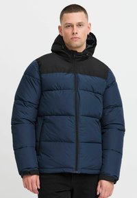 Mørkeblå pufferjakke med sorte accentpaneler, lynlås lukning og hætte. Har et isoleret design med en tekstureret, quiltet overflade.