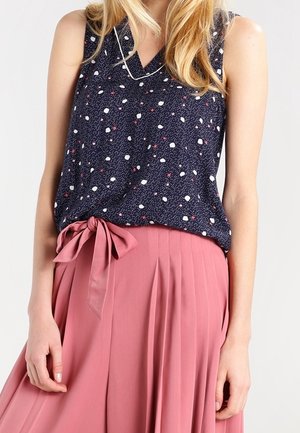 Vrouw die een marineblauwe mouwloze top draagt met witte en rode stippen, gecombineerd met een hooggetailleerde roze geplooide rok die is vastgebonden met een stoffen strik.