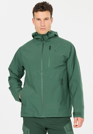 Mann trägt dunkelgrüne, wasserfeste Kapuzenjacke mit Reißverschlusstaschen und verstellbaren Bündchen, steht vor weißem Hintergrund.