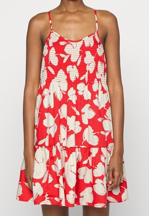 Kobieta ubrana w czerwoną, bezrękawną sukienkę na lato z białym, abstrakcyjnym kwiatowym wzorem i cienkimi ramiączkami, stojąca na jednolitym tle.