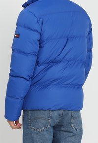 Blauwe pufferjack met gewatteerd ontwerp, hoge kraag en logo-opdruk op de mouw. Heeft een relaxed fit en een gladde, glanzende buitenkant.