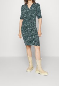 Femme portant une robe verte à fleurs, au genou, avec des manches trois-quarts et des bottes chunky beiges mi-mollet, se tenant sur un sol uni.