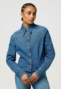 Jeansjakke med knappestilling foran, krage og vestlig stil sømmer. Har blått stoff med mellomvask, lange ermer og trykknapper.