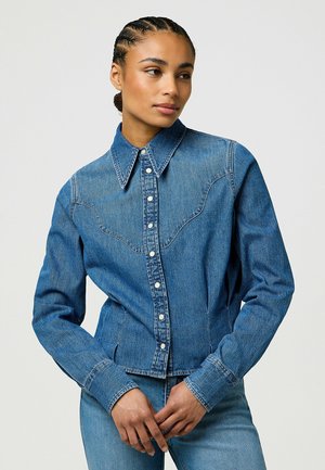 Jeansjacka med knäppning framtill, krage och västerländska sömmar. Har blått tyg med en medelhög tvätt, långa ärmar och tryckknappar.