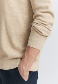 Beige gestrickter Pullover mit strukturierter Oberfläche und gerippten Ärmelbündchen. Die Trägerin hat eine Hand in einer marineblauen Tasche, was den lässigen Sitz des Pullovers zeigt.