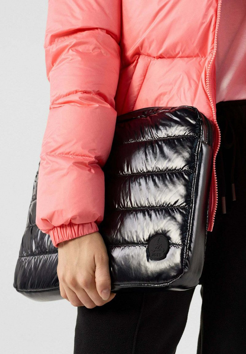 Zwarte puffy tas met een gequiltde textuur en glanzende afwerking, voorzien van een rond logo-patch. Vastgehouden door iemand die een roze pufferjack draagt.