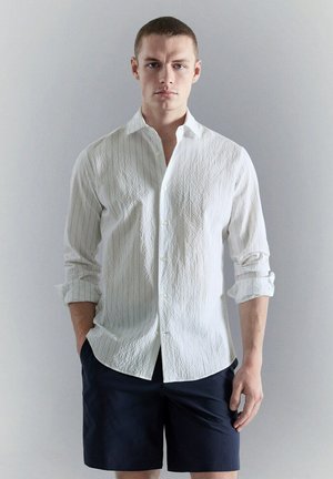 Hombre con pelo corto que lleva una camisa blanca de botones con textura, mangas remangadas y pantalones cortos oscuros, de pie frente a un fondo liso y claro.