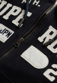 Sudadera con cremallera en color azul marino, con bordados y parches blancos. Cuenta con una cremallera de latón y un cordón con puntas de metal. Tela suave y texturizada.