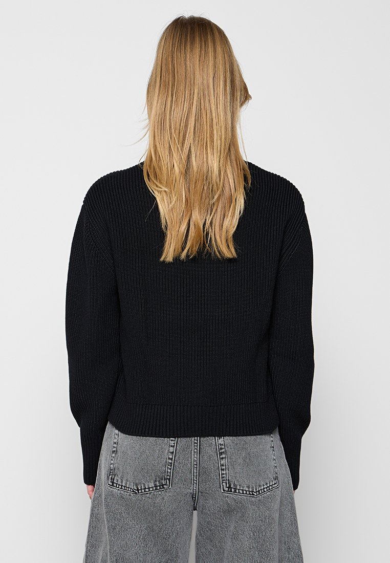 Pull à côtes noir avec un design court et des manches larges, fabriqué dans un tissu texturé. Porté avec un jean taille haute gris clair.