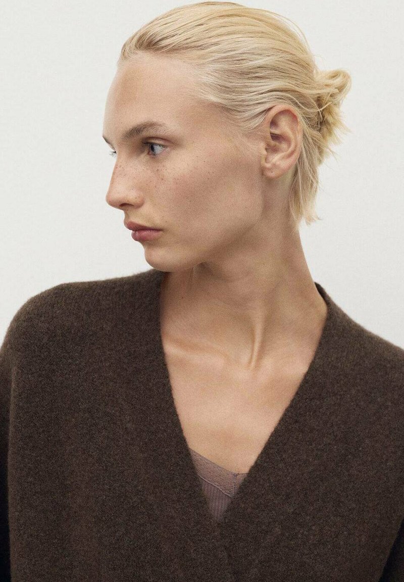 Cardigan en laine brun avec un décolleté en V profond, texture douce et coupe décontractée, porté par-dessus un débardeur beige clair. Le modèle montre un profil latéral.