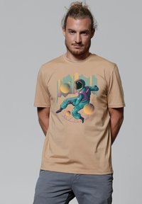 Beige T-shirt met een kleurrijke astronautafbeelding in turquoise en paars, met een gladde textuur en korte mouwen.
