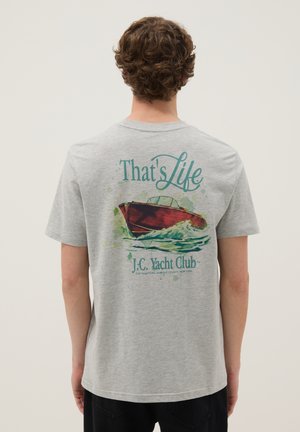 Giovane con capelli ricci che indossa una maglietta grigia con una barca rossa e la scritta "That's Life J.C. Yacht Club" sul retro.