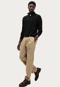 Maglione nero con zip, pantaloni beige plissettati e scarpe marroni con lacci. Il modello posa in modo rilassato su uno sfondo chiaro.
