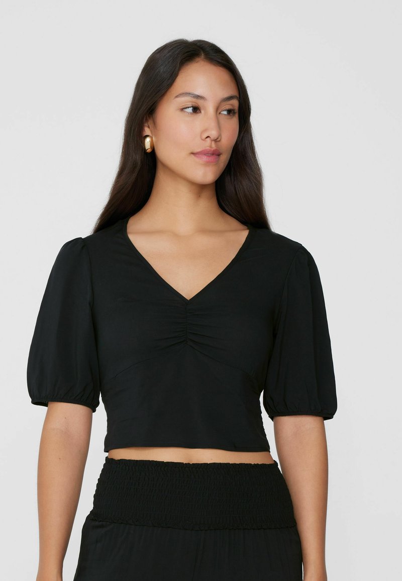 Blusa negra corta con escote en V y detalles fruncidos en la parte delantera. Las mangas cortas abullonadas y una textura suave realzan su diseño.