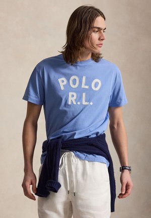 Jonge man met schouderlang haar, draagt een blauw "POLO R.L." T-shirt, witte broek en een marineblauwe trui gestrikt rond zijn taille.