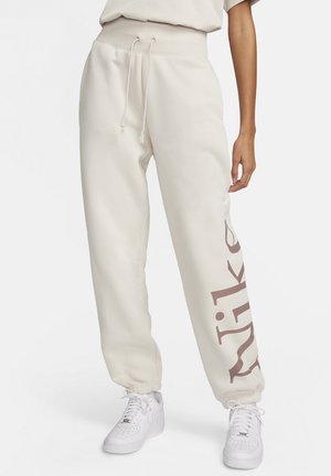 Pantalones de chándal color crema con cintura elástica, cordón y gran logo de "Nike" en marrón claro en la pierna izquierda. Textura suave.