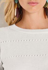 Frau mit großen, mehrfarbigen, hängenden Ohrringen und einem weißen Strickpullover mit dekorativen horizontalen Löchern in der Nähe des Ausschnitts.