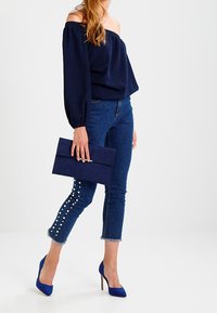 Marinblå off-shoulder topp, blå jeans med pärlprydnader vid vristerna, blå högklackade skor och en marinblå clutch.