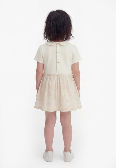 Παιδί με σκούρα μαλλιά που φοράει εκρού φόρεμα κοντομάνικο με κουμπιά πίσω και λευκά αθλητικά παπούτσια, στέκεται με την πλάτη στραμμένη.