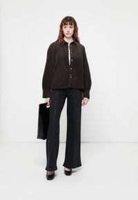 Cămașă oversized din corduroy maroniu cu nasturi aurii, asortată cu blugi negri evazați. Modelul ține o geantă de umăr neagră și poartă tocuri negre.