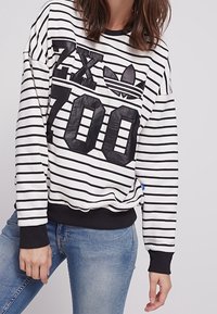 Bílá a černě pruhovaná mikina s velkým černým grafikem "ZX 700" a logem Adidas na přední straně, s žebrovanými manžetami a lemem.