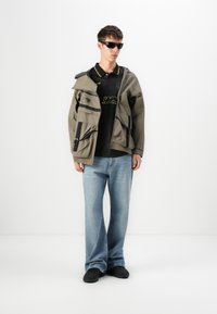 Olivengrønn jakke med flere lommer, svarte detaljer; svart pikéskjorte med gul kant; lyseblå straight-leg jeans; svarte sko.