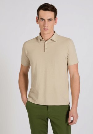 Uomo che indossa una polo a maniche corte beige e pantaloni verde oliva, in piedi con una mano in tasca davanti a uno sfondo neutro.