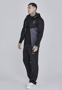 Černý a šedý teplákový kostým s mikinou na zip a kapucí, zúženými tepláky a bílými teniskami; obsahuje loga a má hladkou texturu.