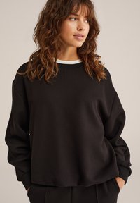 Felpa nera con collo a giro, vestibilità ampia, maniche lunghe e dettaglio bianco al colletto. Tessuto morbido con un design minimalista.