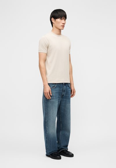 Νέος άνδρας που στέκεται φορώντας μια απλή μπεζ κοντομάνικη μπλούζα, φαρδιά μπλε τζιν και μαύρα παπούτσια, μπροστά σε ένα απλό ανοιχτό φόντο.