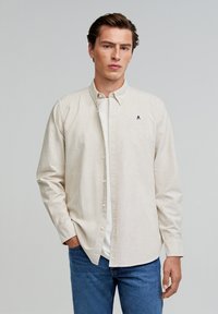 Cremfarbener und beiger gestreifter Hemd mit Kragen, langen Ärmeln und einem kleinen marineblauen Logo auf der Brust, kombiniert mit blauen Jeans.