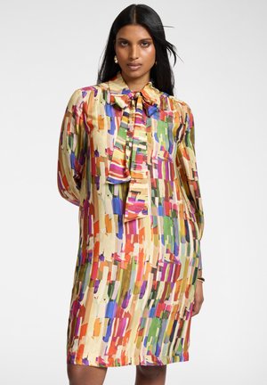 Vrouw met een knielange jurk met een kleurrijk abstract patroon, een strikboord en lange mouwen, staand tegen een witte achtergrond.