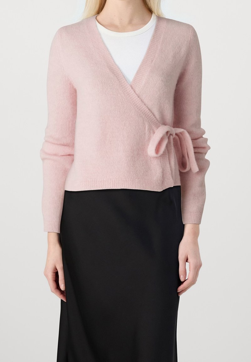 Maglione avvolgente rosa chiaro realizzato in un tessuto morbido, con un dettaglio a cravatta in vita e maniche lunghe aderenti, abbinato a una gonna nera.