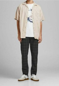 Beżowa koszula z krótkim rękawem na białym t-shircie z grafiką, w połączeniu z ciemnoszarymi spodniami w kratę i białymi trampkami z brązowymi podeszwami.