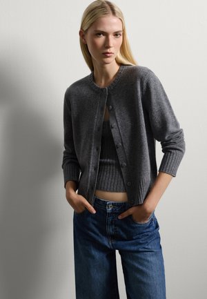 Grau gestrickter Cardigan mit Knopfverschluss, gerippten Bündchen und passendem Tanktop, kombiniert mit hochgeschnittenen blauen Jeans. Sanfte Textur.